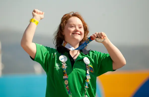 Athlete in green shirt with different pins around the front, holding up their medal